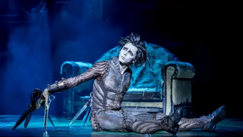 Johan Persson An actor playing Edward Scissorhands sits on stage at the Bristol Hippodrome in his dark costume with large pairs of scissors attached to both of his hands and a faded, tatty sofa behind him. The stage is illuminated with a blue/white light.