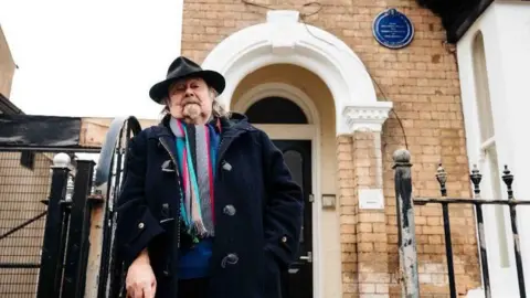 A man is stood outside an open black gate which leads to the front door of a property with an archway. He is wearing a thick black coat, black hat and colourful scarf.