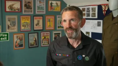 A man with a black shirt and badges looks into the camera.