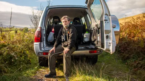 Dafydd Hughes holds a whip and looks into the camera. He is sat connected  the ledge of his conveyance  with the footwear  doorway  open. 