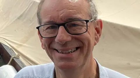 Simon Coupland Simon Coupland, who is clean shaven, wears black-framed glasses, a light grey shirt and has short grey hair. He is smiling broadly.