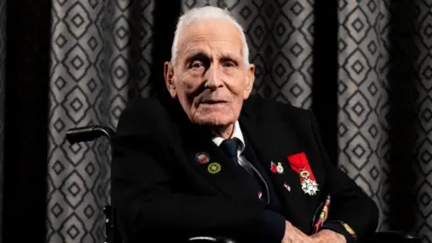 PA Media D-Day veteran John Dennett, from Merseyside, pictured in 2024. He is wearing a dark suit with medals on it and sitting in a wheelchair. The background is geometric grey curtains.