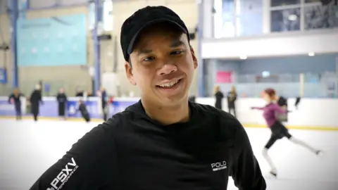 Hanyu Wong. He is wearing a black baseball cap and a long sleeve black top. He is smiling at the camera. Behind him is an ice rink with several people skating.