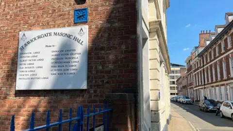 LDRS The side of a large, off-white building on a residential street. A sign affixed to the side of the building reads: St Saviourgate Masonic Hall.