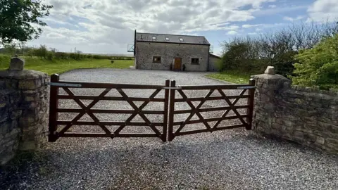 Stone yard with a gate. Behind the gate is a grey brick cottage with two small windows and a brown front door. The roof of the cottage has three small windows. There is two balconies either end of the property.