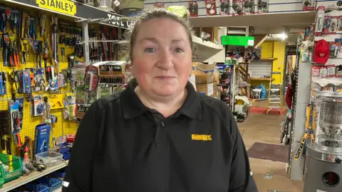 Jennifer Murray wears a black polo top with a yellow logo on the chest. She stands in the middle of the hardware store that she works in.