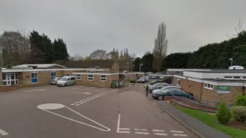 Google A car park with one-storey buildings on either side that look like an infant school. There are a few cars parked around the edges and a man wearing a high vis vest walks towards the camera, at a distance. There are trees and hedges behind the buildings. 