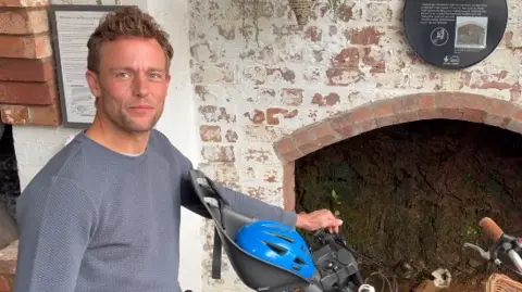 A man with short dusty blonde hair is wearing a blue long-sleeve top while sitting on a bike with a child seat on the front. A blue helmet is in the seat