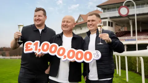 Anthony Devlin Paul with his two sons who are holding glasses of champagne and standing either side of him smiling at the camera. Paul holds a large cut-out with the figure £1,000,000 in orange, and the National Lottery logo. They stand on the racecourse grass. 
