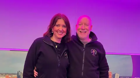 A woman with dark hair hugging a bald man man in a radio studio with a purple backdrop 