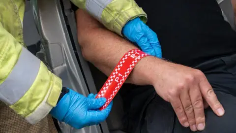 Northamptonshire Fire and Rescue Service A triage band, in red, with P1, written on it, being put on a mans hand, by a firefighter. The man wearing dark clothing is sat in a car.
