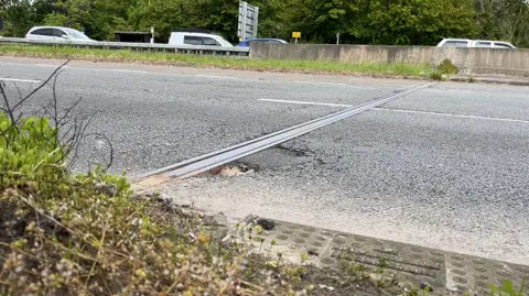 A metal seam in a dual carriageway bridge, which has some cracks on either side. Traffic can be seen queueing on the other side of the dual carriageway. There is a concrete central reservation between both carriageways. There are small grass verges on either side and trees can be seen bordering the other carriageway.