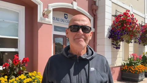 A man wearing a black hoodie and black sunglasses stands by some hanging baskets full of flowers outside a building in the sunshine.