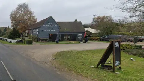 Google Single-story pub painted grey. The words "The Rusty Gun" and "Restaurant" are visible in white lettering. A pub A-frame is visible in the foreground, alongside a driveway edged in grass. A road passes the pub to the left side.
