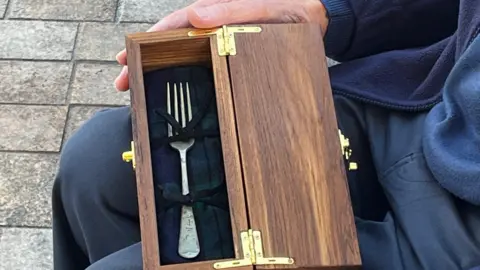 Kevin Shoesmith/BBC A silver-coloured fork in a brown wooden presentation box with gold-coloured hinges. It rests on David Balfour's knees.