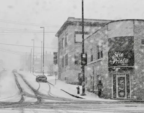 Leonardo Fugoso Jr A black and white image of a road junction covered in snow. There is a shop on the right called 'Skin Prints' and cars driving up a snow-covered road on the left. A man is walking down the steep road, near the shop.