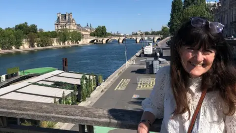 A woman with dark hair stands smiling in front of a river with a beautiful, large building on the other side of the water. She is wearing a white top and has her glasses on her head.