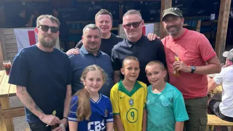 Paul Styles Wayne Rooney at a pub garden with four men and three children all gathered near him and smiling. The kids are all wearing football shirts.