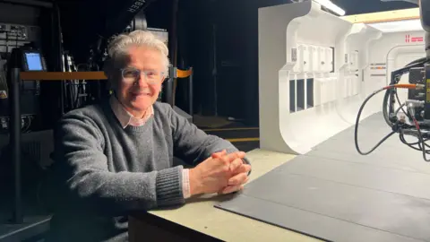John Lee John Lee is smiling, with silver hair and glasses, while stood near a model of a spaceship corridor set from Rogue One.