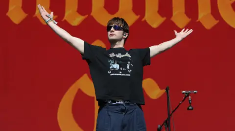 The lead singer of Fontaines D.C., Grian Chatten, pictured performing standing with his arms in the air and sunglasses on.