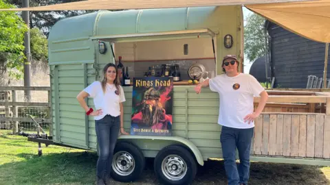 Darren Rozier/BBC A woman and a man wearing white T-shirts with the King's Head logo on them. They are standing in front of a mobile bar and either side of a large poster of The King's Head logo, which depicts a Viking-type character under the words King's Head.