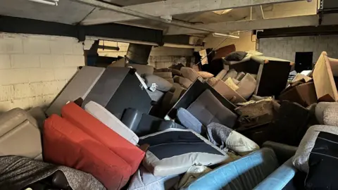 Hewer-White Trust Furniture including sofas and chairs lie piled up in a disorganised manner inside a white-walled industrial unit. 