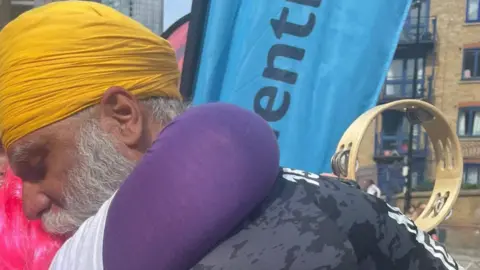 Manny Singh Kang He is hugging a woman with pink hair who is wearing purple clothing on an arm. Part of a blue banner is visible behind them.