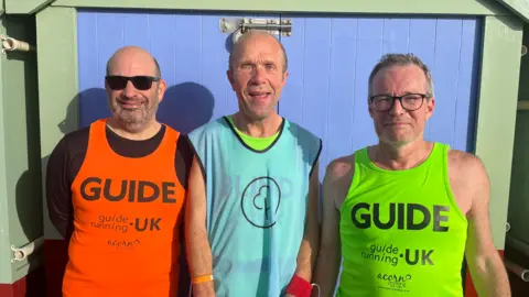 Keith Turner (middle) is joined by two guide runners standing either side of him. Keith is wearing a luminous t-shirt reading "BLIND", branded by Guide Running UK. This sits behind a transparent Parkrun volunteer pacer vest, which he is wearing on top. His guide runners are wearing similar Guide Running UK vests but with the label "GUIDE". The trio are standing shoulder-to-shoulder in front of a beach hut on Hove Lawns seafront.