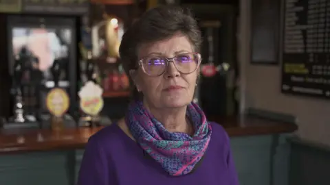 A woman in a purple top, with purple, pink and turquoise snake-skin print scarf. She has short hair and glasses and is looking at the camera with a pensive expression. Behind her a bar and beer taps can be seen.