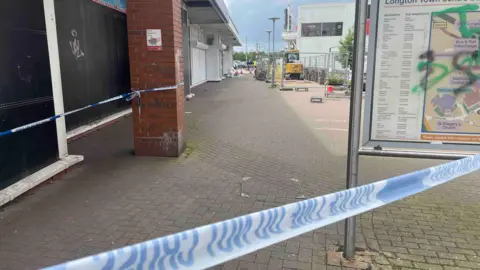 A blue and white police cordon around an area which has a brick pavement and a sign with some graffiti on it. 