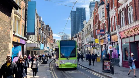 Getty Images A green tram on the street in a built up area of London. There are pedestrians on either side of the street. Tower blocks are visible in the background. 