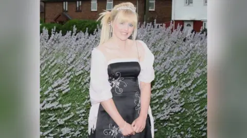 Family photo Abbigail Smith wears a black and silver prom dress with a white shawl over the top. She has blonde hair which is tied at the back, a silver tiara and silver jewellery. She is standing in front of a bush and is smiling at the camera.