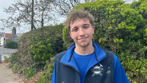 Ben Armitage - A man with brown curly hair, looking at the camera. He's wearing a blue jacket with a blue shirt underneath, there is a paint stain on the left lapel. 