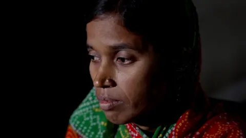 Siraj Ali Lalmunni Devi posa em um quarto escuro em um saree multicolorido. Ela perdeu o marido devido ao consumo de álcool tóxico em 2022. 