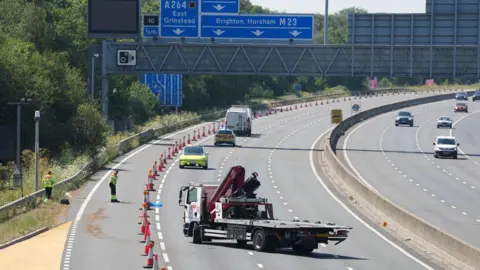 Police and recovery vehicles are in a coned-off section of the motorway. A flatbed recovery truck is parked in the closed lanes while two highway workers in hi-vis clothing stand beside the hard shoulder. Overhead blue motorway signs point to Crawley, East Grinstead, Brighton and Horsham.