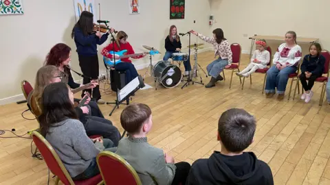 Young musicians sit in a circle with guitars and drums as they rehearse 'Home'