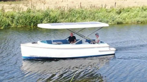 Tim Knox and his wife Sue travelling on the boat. It is a small white vessel with solar paneling on its roof.