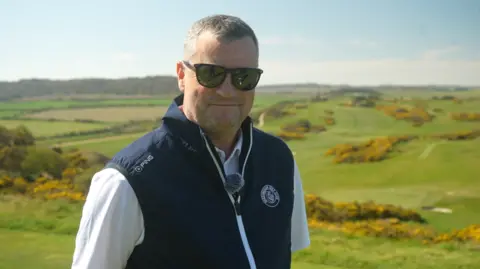 Paul Innocent has cropped grey hair and is wearing sunglasses, a white shirt and navy jacket. He is smiling and is standing on the fairway.