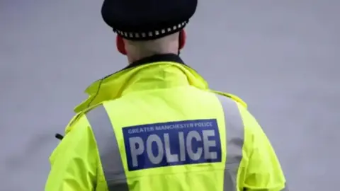 A police officer faces away from the camera. He wears a high vis jacket with Greater Manchester Police on the back.