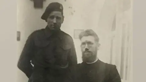 John Lloyd A black and white photo of Jack Lloyd, standing in Army uniform with a beret on his head. Sitting to his left is a bearded young Brussels' priest in black with a white dog collar.