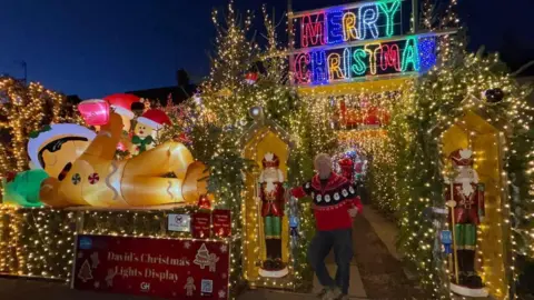 Sue Ryder David, a man, wearing a red Christmas jumper and blue jeans, standing in front of his Christmas display with lights and inflatable snowman and nutcrackers and a merry Christmas sign lit up in multi-colours. 