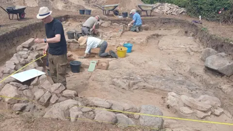 Four people excavate a shallow trench with stones visible near the surface.