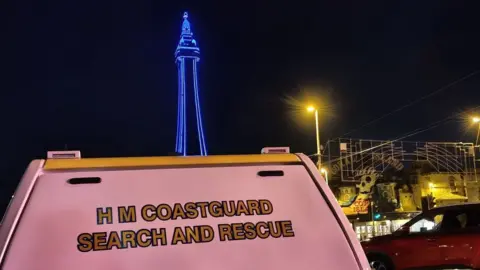 An image at night showing the top of a white coastguard van that says "HM Coastguard Search and Rescue" below Blackpool Tower outlined in blue lights and a view of the lit-up pier to the right.