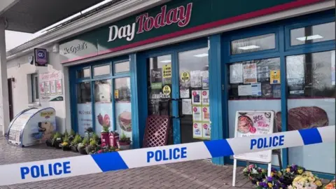 A police cordon can be seen outside a shop. 
A number of plants can be seen outside it and the words: 'Day Today' are visible on a sign above the shopfront.