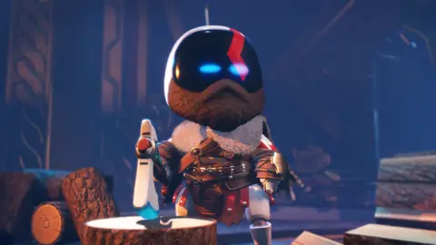 Sony Pictures Astro Bot - a white, cute robot with light blue eyes - dressed like Kratos, the protagonist from God of War. Astro has a beard, and wears a leather jacket with many buckles and a fur collar. Astro stands next to a cut log, holding a large ax and surrounded by trees.