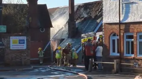 A firefighter aims a jet of water at the steeply-pitched roof of Emsworth Community Centre. At least eight other people, including firefighters and members of the public, stand nearby.