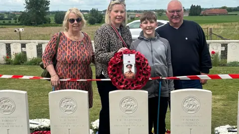 Whitaker family Four people stand behind war graves, there is plastic tape around the graves and flowers and little crosses at the foot of the headstones. 