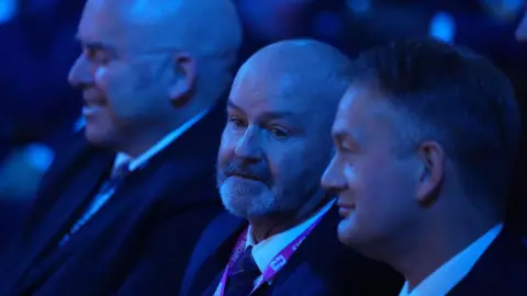 Reuters Steve Clarke, a bald man with beard sitting next to other men at the world up draw. There is a blue light on them.