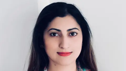 Supplied Rahat Khan smiles at the camera against a white background. She has long dark hair and is wearing red lipstick. 