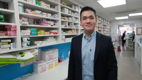 Un farmacéutico se encuentra junto a estantes blancos llenos de medicamentos en la trastienda de una farmacia. Tiene el pelo corto y negro y viste una elegante chaqueta negra sobre una camisa azul claro. Al fondo, se ve a una mujer de pie en la entrada de la farmacia.